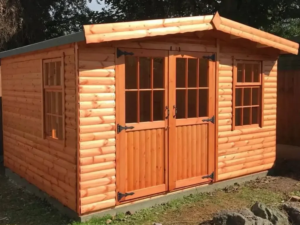An inviting shed log cabin with a warm finish, decorative window boxes, and large glass-paned doors, situated on a modern grey paver patio.