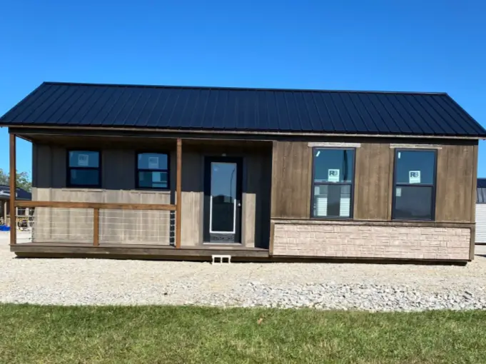 A 16x32 ranch tiny home shell with dark wood siding, a black metal roof, and a small porch, sitting on a gravel lot.