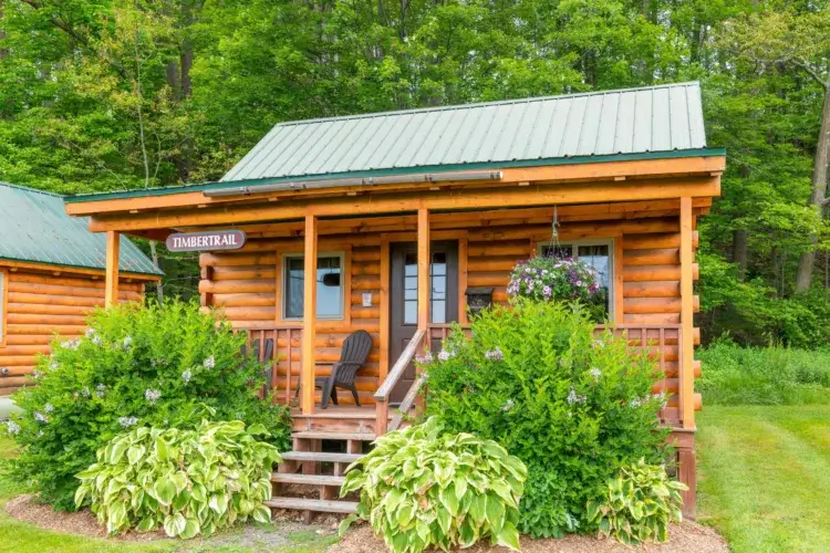 The Timbertrail small log cabin kit with lush landscaping and a hanging flower basket on the porch.