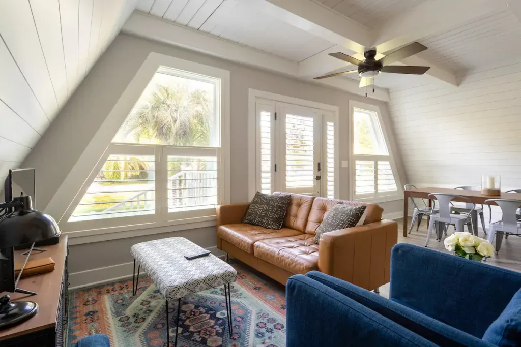 A-frame living area with white shiplap walls and ceiling, a tan leather sofa, a blue armchair, and windows with white plantation shutters.