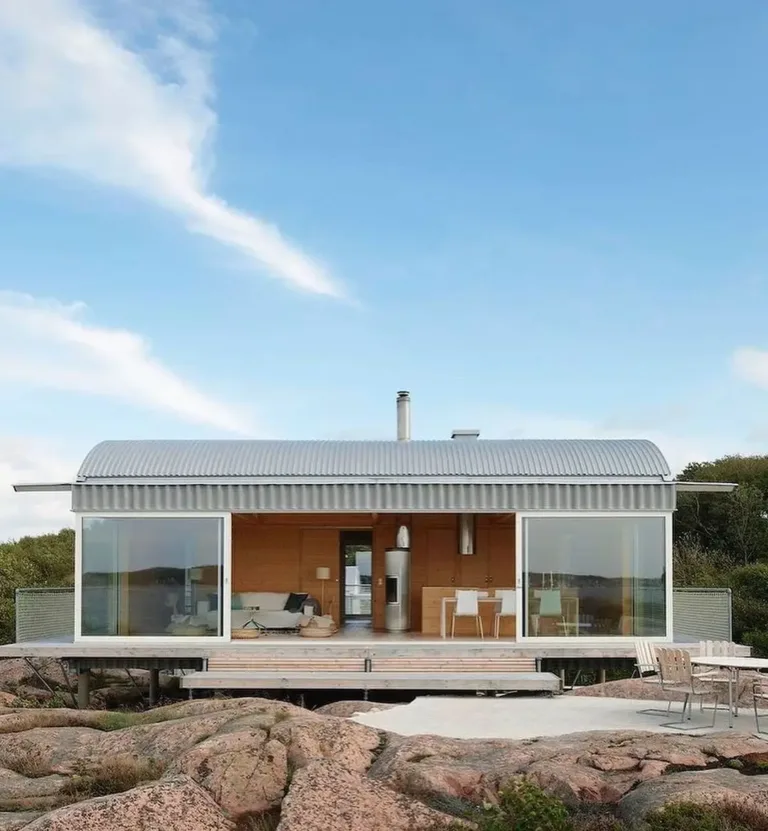 Modern wooden cabin on stilts with a curved roof and wrap-around balcony, overlooking a Swedish fjord.