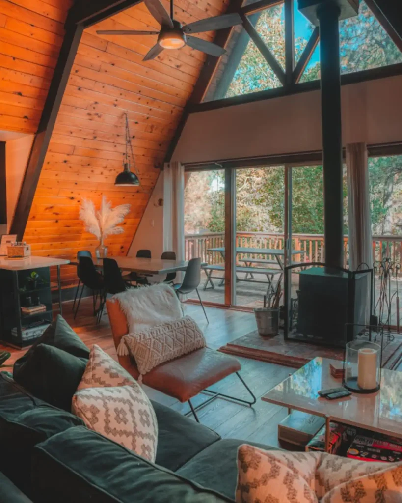 Warmly lit A-frame cabin living room with extensive wood paneling, a black wood-burning stove, a dark sofa with patterned pillows, and a dining area in the background.