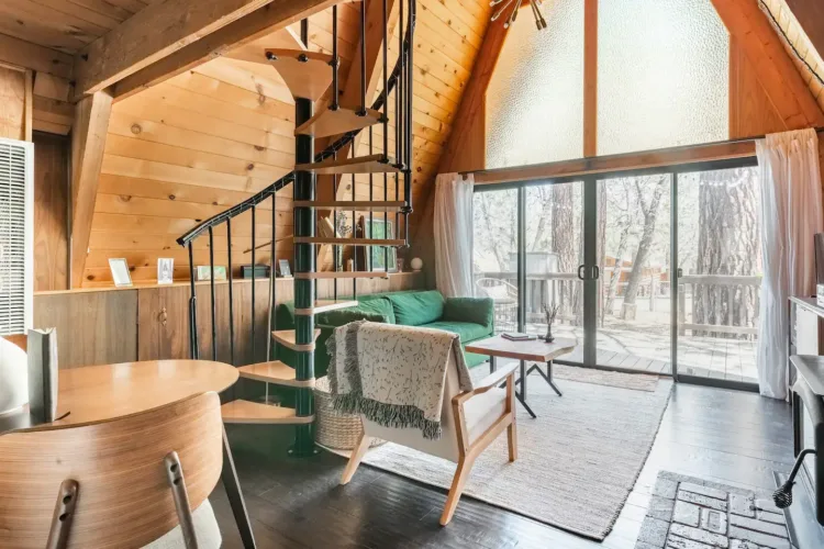 Modern rustic A-frame house interior ('Whispering Waters') featuring a black spiral staircase and light wood paneling.