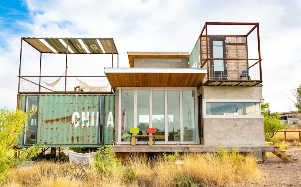 The Hermosa Container Home, an eclectic shipping container home design in Marfa, TX, featuring weathered green containers, multiple decks, and a rustic desert aesthetic.