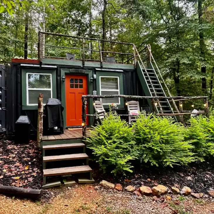Design of the 'IN THE WOODS' shipping container home near Cheaha State Park, AL, with a dark exterior, red door, rooftop deck, and fire pit in a colorful autumn forest.