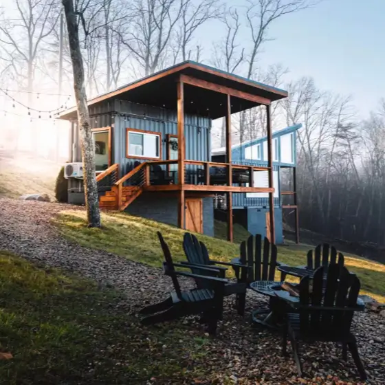 Modern shipping container home design of The Belvedere Box in Rising Fawn, GA, featuring a dark grey exterior, covered wooden porch, and a fire pit in a serene woodland setting.