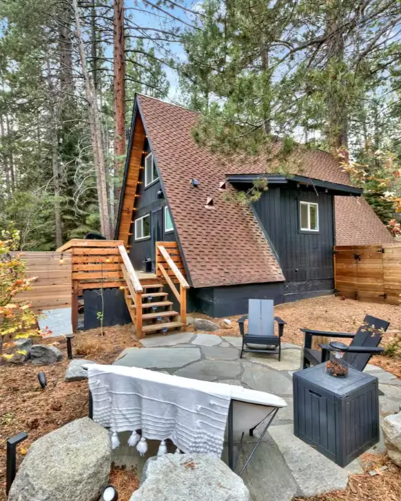 The stylish, modern exterior of the KÜLHAUS Tahoe A-frame cabin, featuring a stone patio with outdoor seating nestled among the trees in South Lake Tahoe.