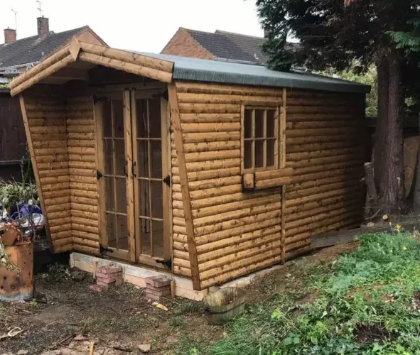 A newly-built, unstained pine shed log cabin from a kit, featuring a glass-paned door and a window with matching wooden shutters.