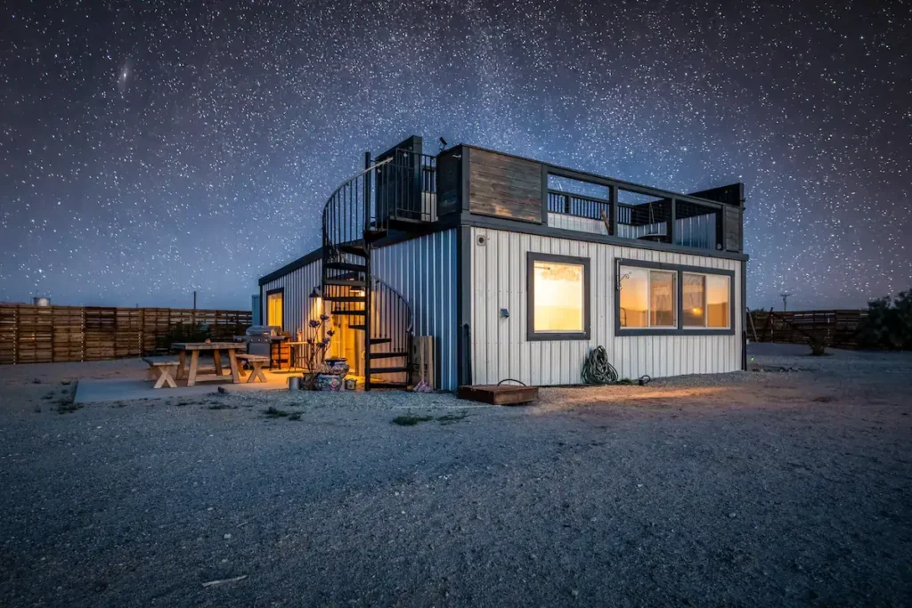 Design of The Glow House in Twentynine Palms, a light-colored shipping container home with a spiral staircase leading to a rooftop deck, optimized for desert stargazing.
