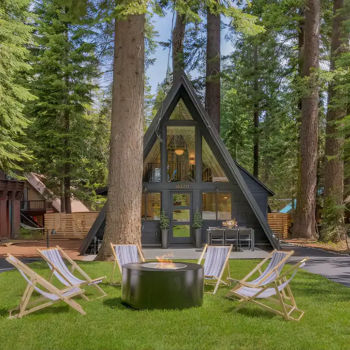 The inviting front yard of the Little Owl Tahoe A-frame cabin in Truckee, featuring canvas chairs circled around a modern fire pit on a lush green lawn.