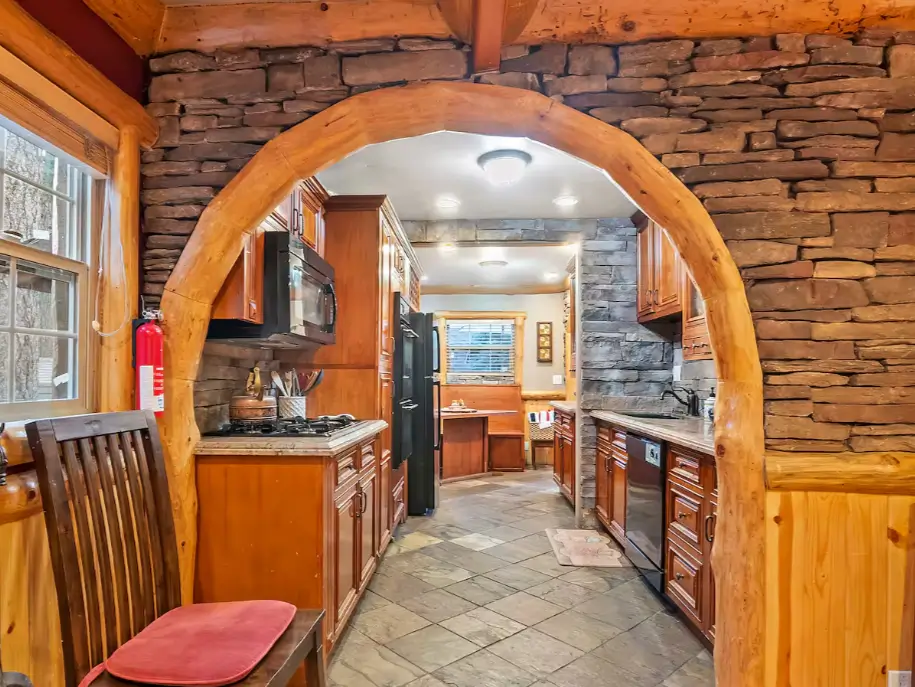 A stunning handcrafted log and stone archway leading into the rustic gourmet kitchen of a Big Bear cabin.