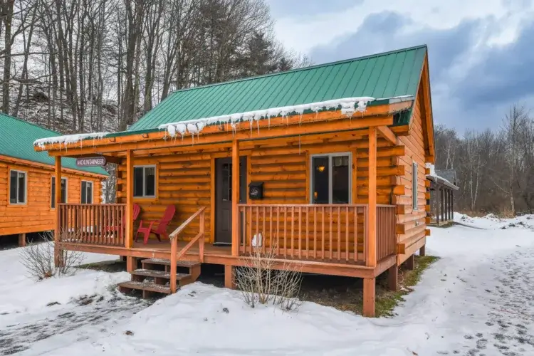 The Mountaineer log cabin kit in a snowy landscape, featuring a full-length porch with red chairs.