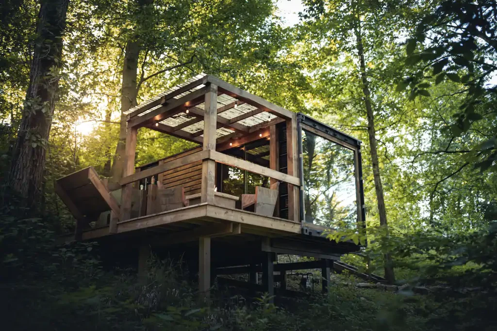 Design of Forest Haven - Otium in Millersburg, OH, a modern shipping container tiny home with an elevated wooden deck and translucent pergola, creating a treehouse feel in a sun-dappled forest.