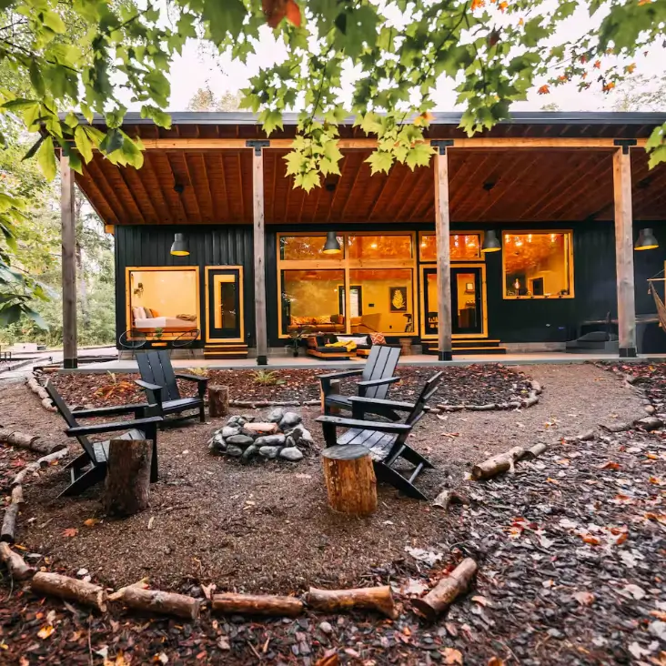 Modern ranch-style shipping container home design of The Wren in Logan, OH, with a dark exterior, expansive glass walls, a deep covered patio, and a cozy forest fire pit.