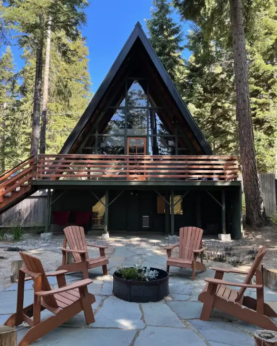 The classic exterior of the Modern Mid-Century Tahoe A-frame in Carnelian Bay, showing its stone patio with Adirondack chairs gathered around a fire pit, all set beneath towering pine trees on the North Shore.