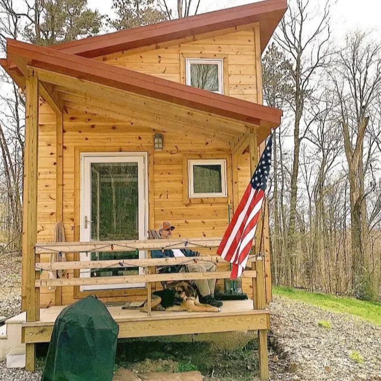 A man and his dog relax on the porch of a rustic wooden tiny house in an Ohio forest, representing the tiny living lifestyle.