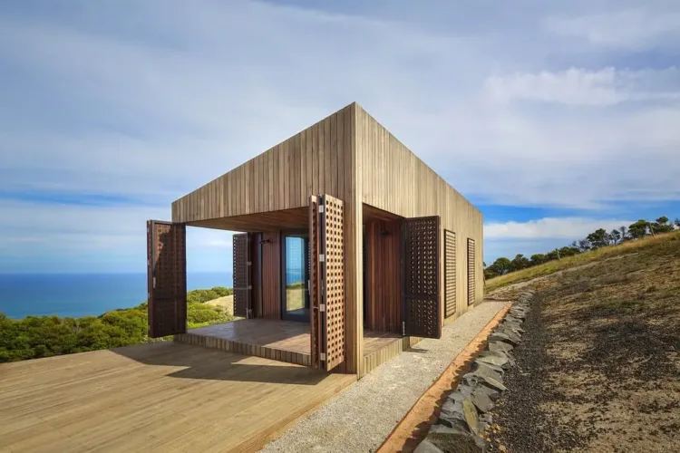 Small, eco-friendly cabin exterior on a wild Australian coastline, featuring light-colored wood and clean lines.