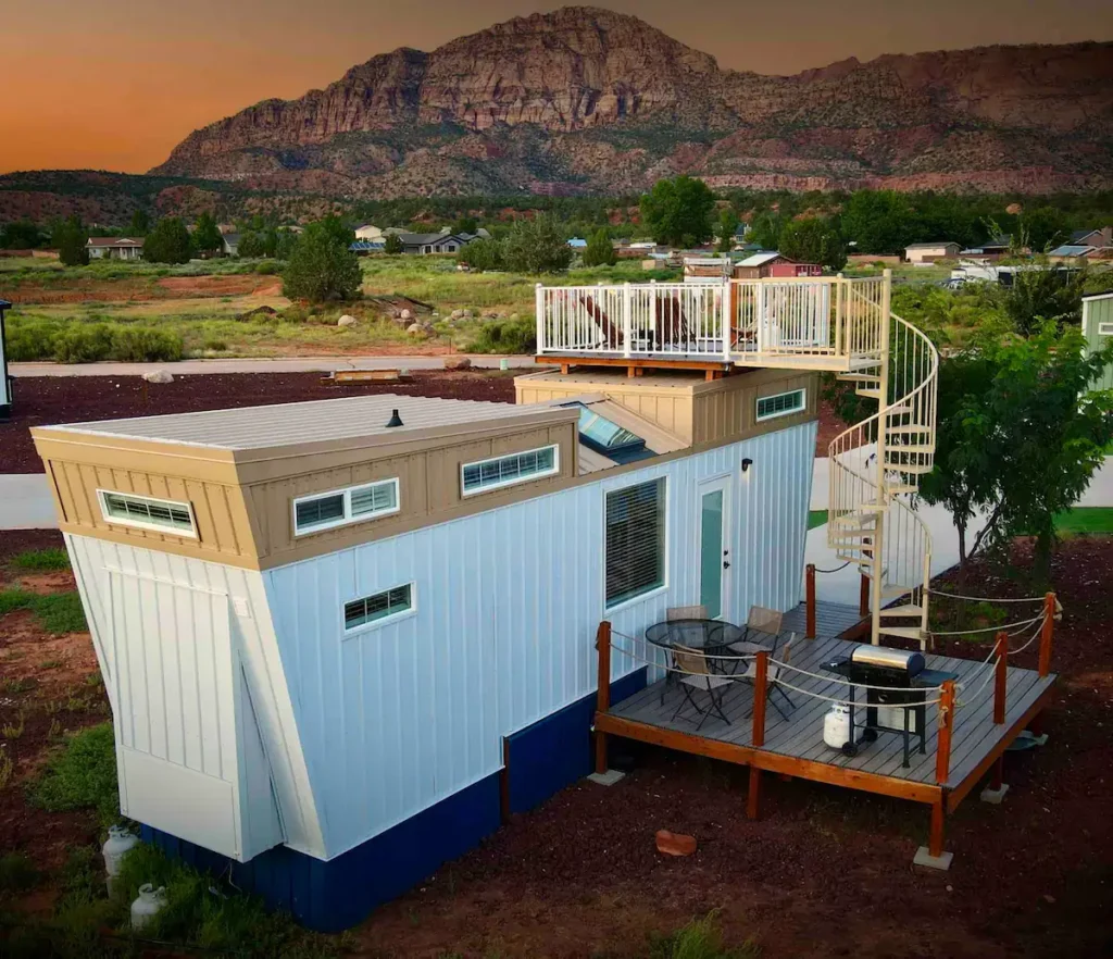 Unique shipping container style home design of The Starboard in Apple Valley, UT, with an angled white facade, tan upper section, rooftop deck with spiral staircase, and dramatic desert mountain views.