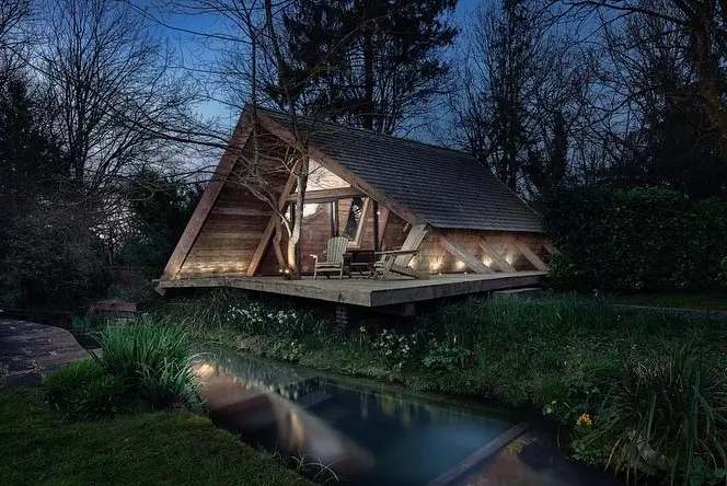 The Climber's Cabin in the UK, a cozy A-frame wooden cabin by a stream, warmly lit at dusk, epitomizing hygge and rustic Scandinavian charm.