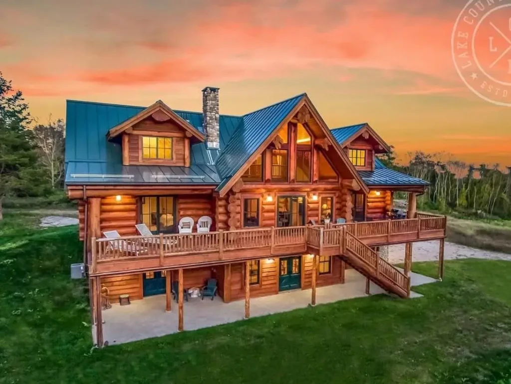 A beautiful two-story handcrafted log home with a large deck and green metal roof, glowing under a warm sunset.