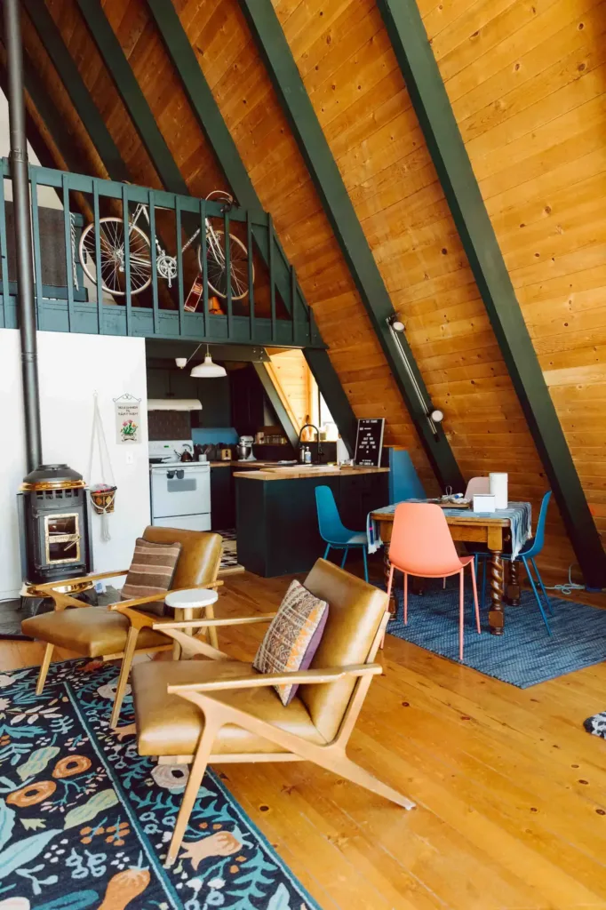 Eclectic A-frame cabin interior design with wood paneling, contrasting dark green beams, and colorful chairs.