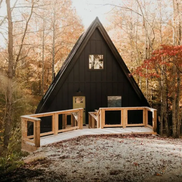 Striking black A-frame cabin with a welcoming wooden ramp and deck, nestled amidst vibrant autumn foliage.