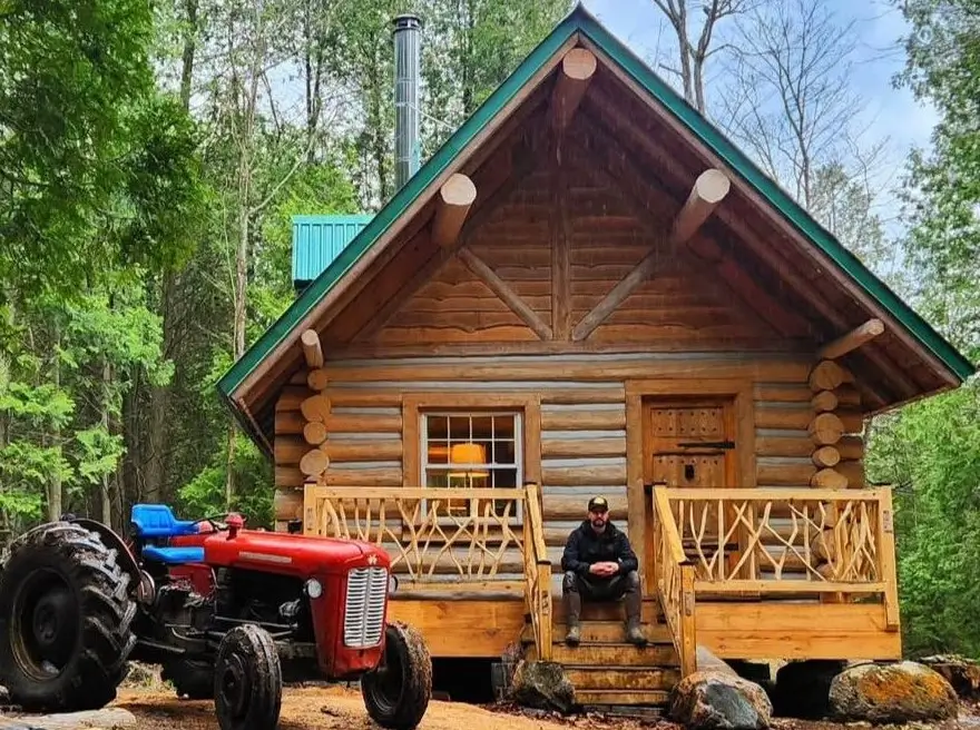 A cozy, handcrafted full-round log cabin nestled in the woods with a man sitting on the porch steps next to a red tractor.