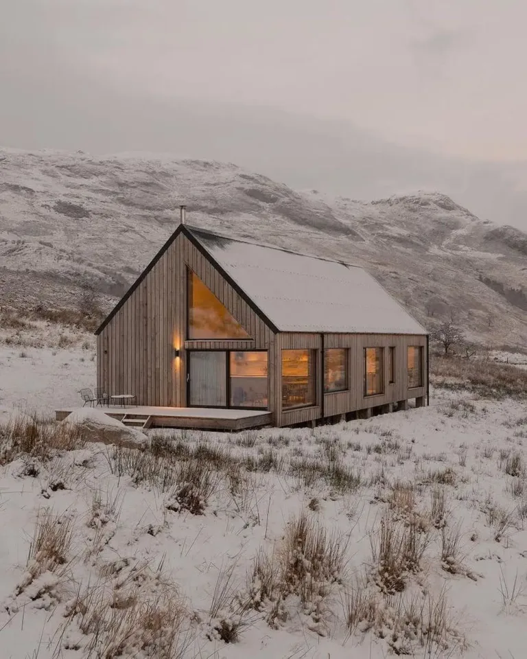 Modern Scandinavian cabin exterior in a snowy landscape, featuring light wood cladding and large windows glowing warmly.