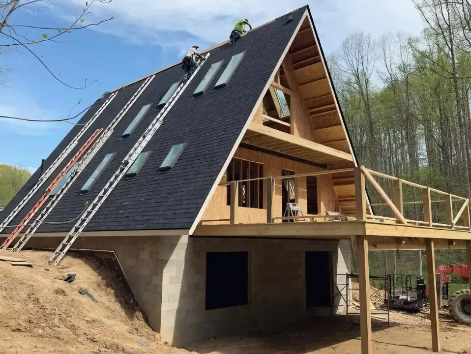 Exterior of an A-frame house nearing completion, featuring a shingled roof with multiple skylights, exposed gable framing, and a wooden deck structure, part of the A-frame building process.