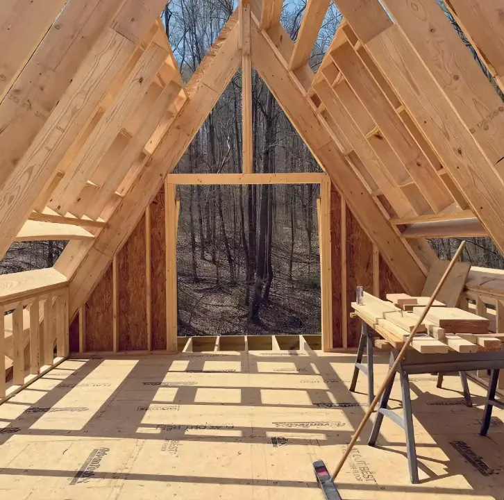 Interior perspective of an A-frame cabin during construction, showcasing exposed wooden roof rafters, subfloor, and a large framed opening offering a forest view, illustrating A-frame spatial characteristics.