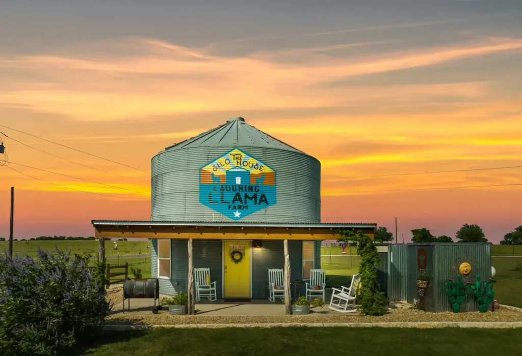 The Silo House at Laughing Llama Farm, a unique grain silo rental in Texas, shown at sunset with its bright yellow door and colorful logo.