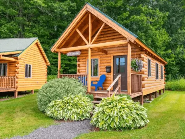 A beautiful Creekside small log cabin kit with a welcoming front porch and lush summer landscaping, featured in a guide to the best cabin kits.