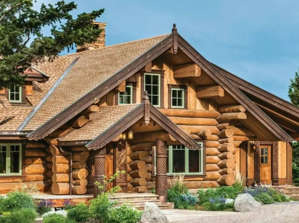 A beautiful handcrafted log cabin featuring full-round logs, intricate decorative woodwork on the porch posts and gables, and surrounded by lush landscaping.