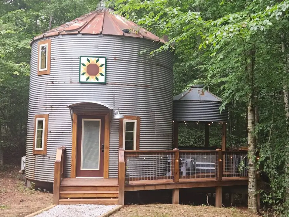 A rustic corrugated metal silo rental, decorated with a sunflower quilt block, nestled in a peaceful wooded area with a small deck.