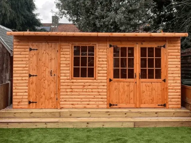 A beautiful shed log cabin with a warm wood stain and double glass doors, sitting on a modern wooden deck in a backyard.