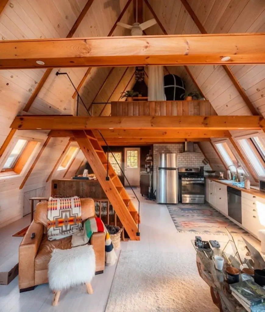 Bright and airy simple A-frame house interior with light wood paneling, exposed beams, a cozy living area with leather armchair, open kitchen, and wooden ladder leading to a loft.