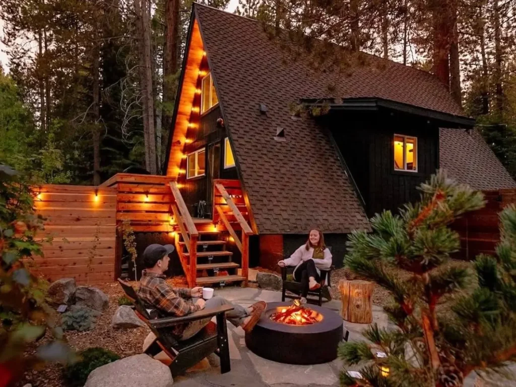 A couple enjoying a cozy evening by a fire pit outside a stylish Tahoe A-frame cabin, with warm string lights illuminating the forest setting at dusk.
