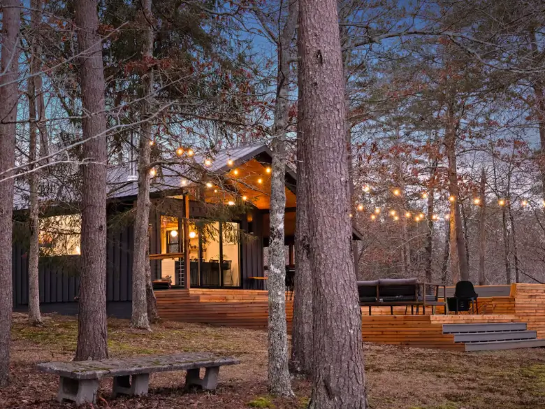 A view of The Coalmont cabin through the trees, with a rustic stone bench in the foreground and warm string lights glowing on the deck.