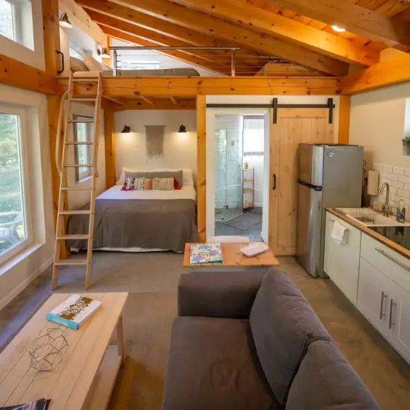 A wide view of the 2 bedroom tiny house interior, showing the open floor plan that connects the living area, kitchen, and the main floor bedroom with a loft above.