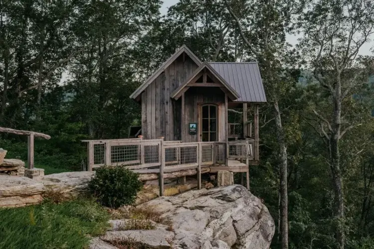 A closer view of the rustic Raven Rock Cabin perched on its cliffside foundation, with a wooden deck and wire railing.