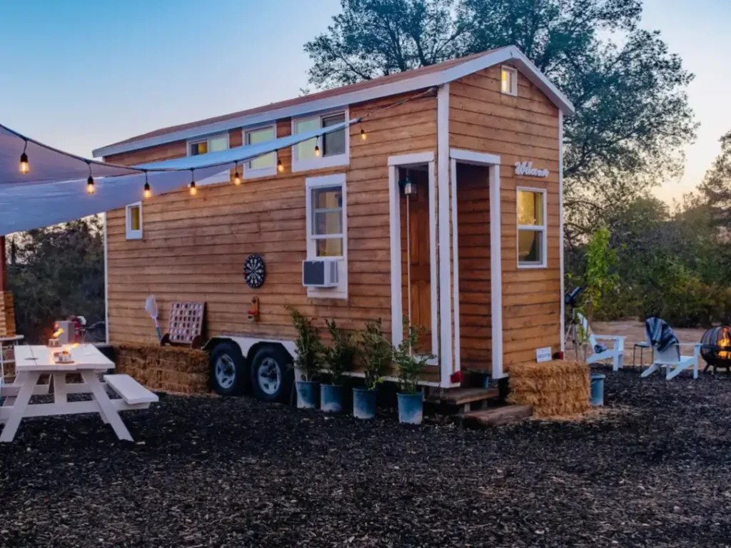 A rustic 200 sq ft tiny house on wheels with natural wood siding, parked in a tranquil setting at dusk with string lights.