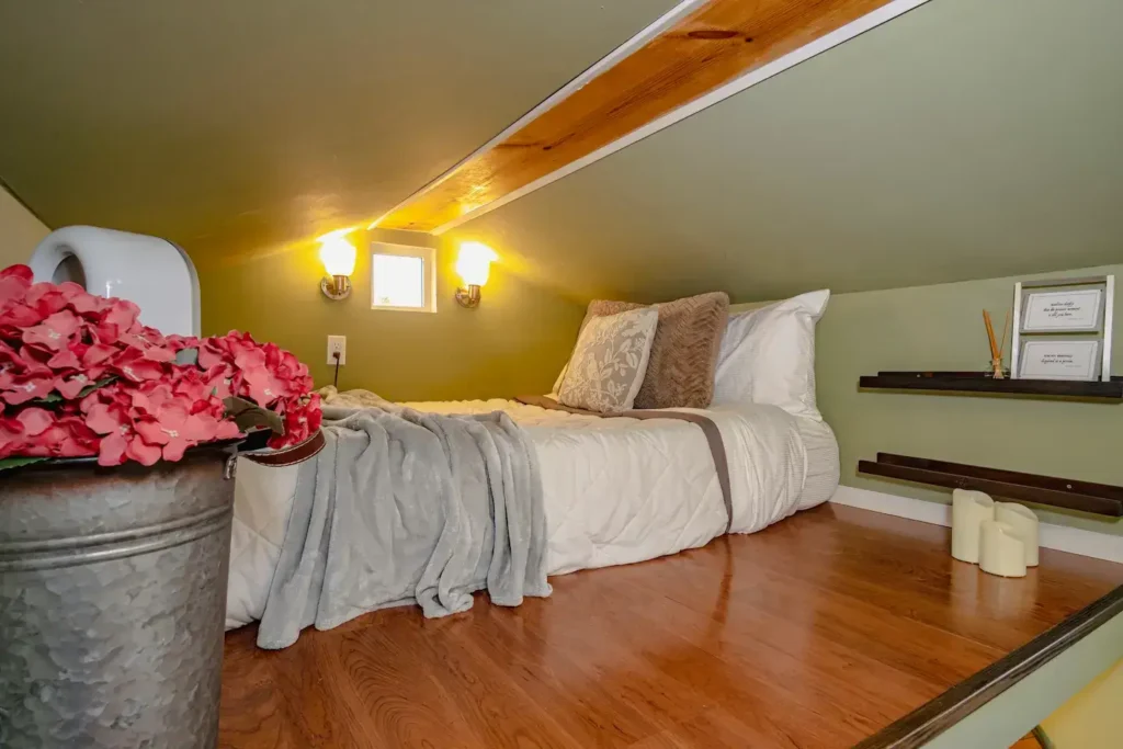 A close-up of a cozy sleeping loft in a 200 sq ft tiny house, with a neatly made bed, pillows, and wall-mounted sconce lighting.