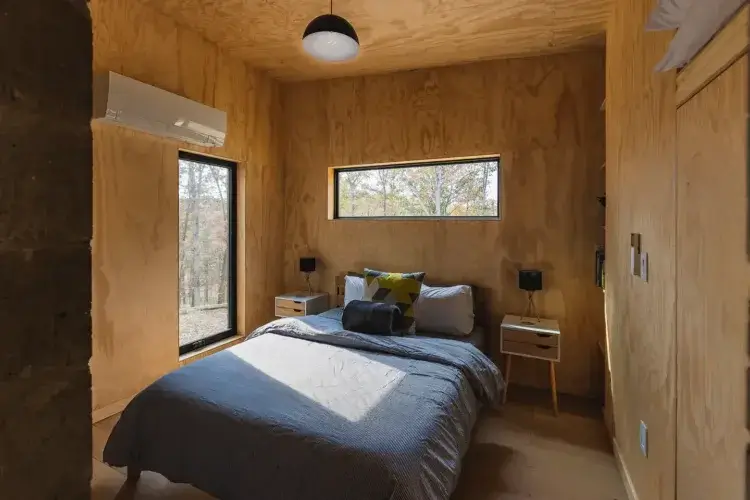 The cozy, minimalist bedroom in the Plahaus Cabin, with a queen-sized bed, simple nightstands, and windows looking out into the woods.