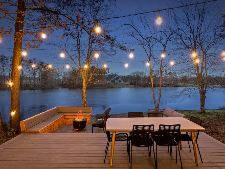 The outdoor dining area on the deck of The Coalmont cabin at dusk, featuring a fire pit, built-in seating, and string lights overlooking the lake.