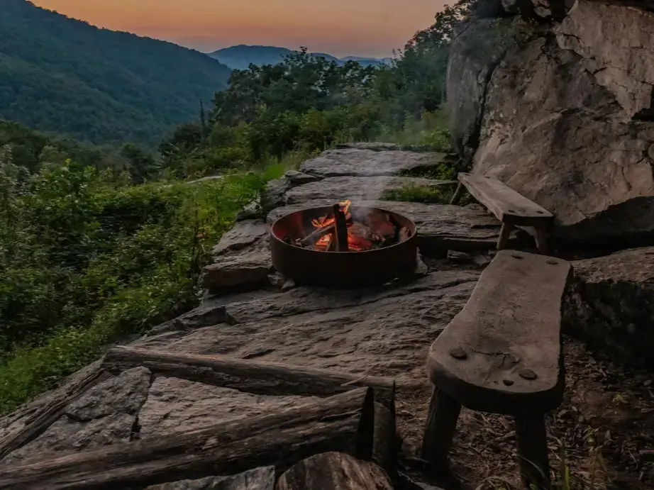 A cozy fire pit with a crackling fire on a stone ledge at the Raven Rock Cabin, with rustic benches overlooking a mountain valley at sunset.