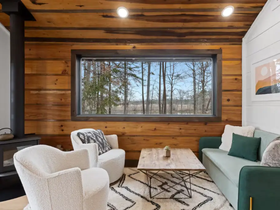 The cozy living room interior of The Coalmont cabin, featuring modern furniture, a wood stove, and a large picture window with views of the surrounding nature.