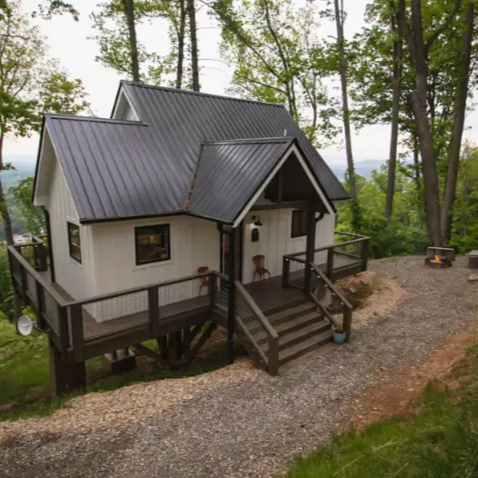 The entrance to the modern farmhouse treehouse in Asheville, showing the wraparound deck, gravel pathway, and a cozy fire pit area for evening relaxation.