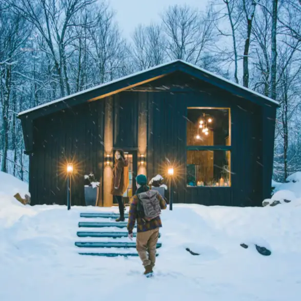 A couple arriving at the warmly lit Birch Falls Spa Cabin for a cozy winter retreat, with snow falling gently around them.