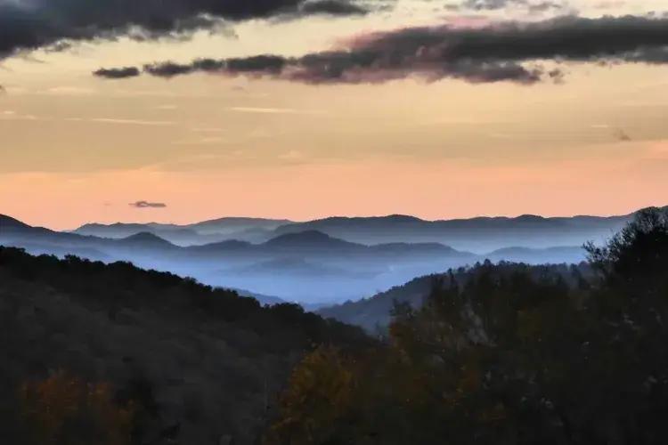 Layers of rolling mountains fade into the distance, with fog settling in the valleys during a colorful sunset, as seen from the Raven Rock Cabin.