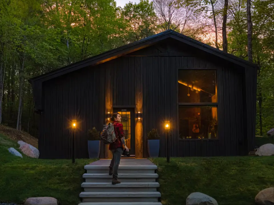 A person with a backpack arriving at the entrance of the modern, black Birch Falls Spa Cabin at dusk, ready for a luxury wellness retreat.